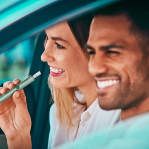 A man driving a car and a woman sitting in the passenger seat holding an IQOS device.