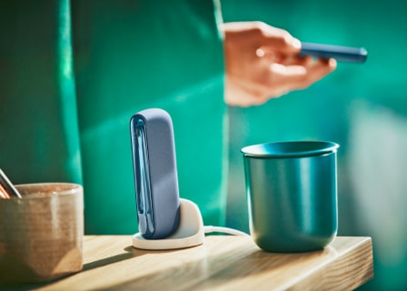  man holds an IQOS ILUMA mid azure blue device, with a holder and cup on the table.