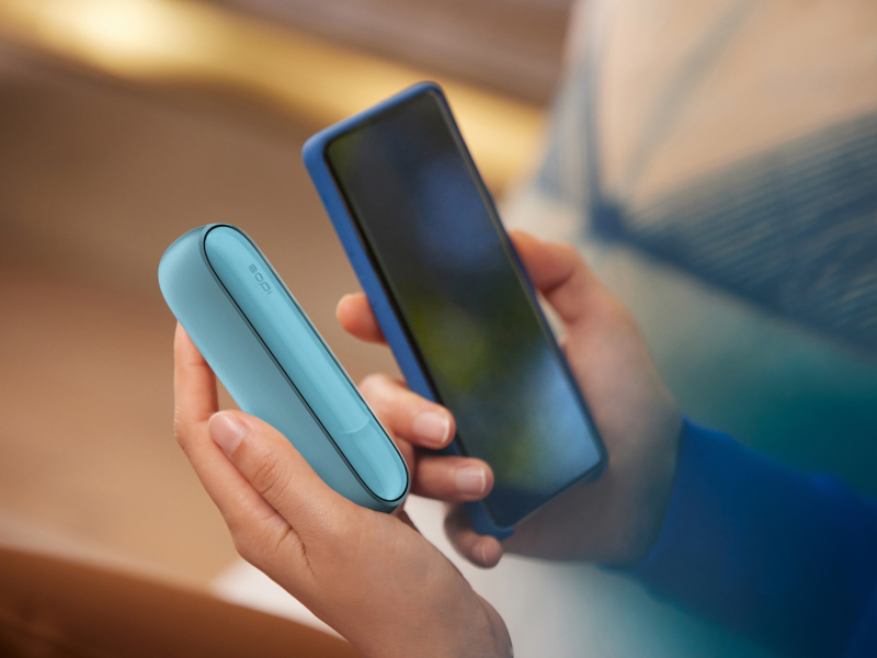Man holding IQOS Originals Duo device in Turquoise in one hand and a mobile phone in the other hand.