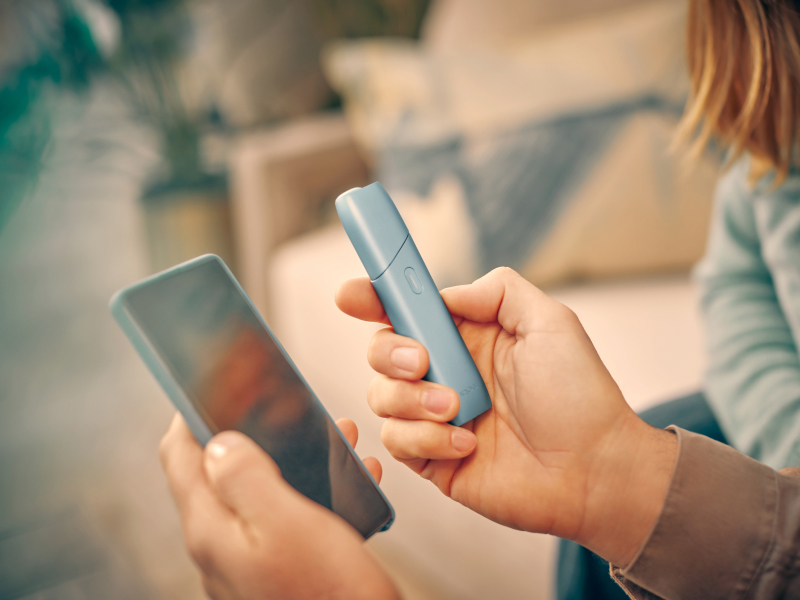 Hands holding a mobile phone and IQOS Originals One device, logging into myIqos to get the most of the heated tobacco device.