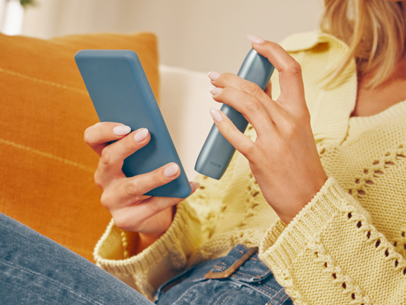 Hands holding a mobile phone and IQOS Originals One device, downloading the app to get a full professionnal maintenance.