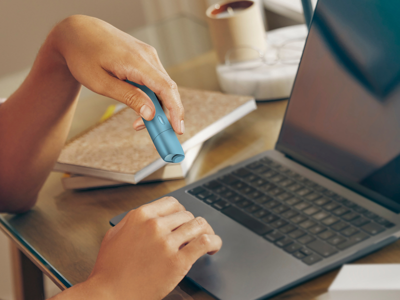 Hands typing on laptop while holding IQOS Originals ONE heated tobacco device.