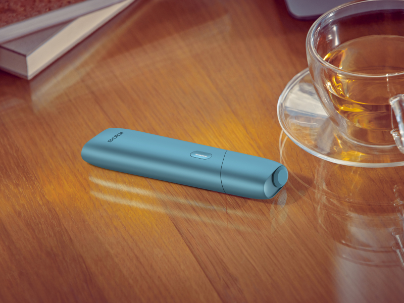 Turquoise IQOS Originals One device on a desk next to a tea cup.