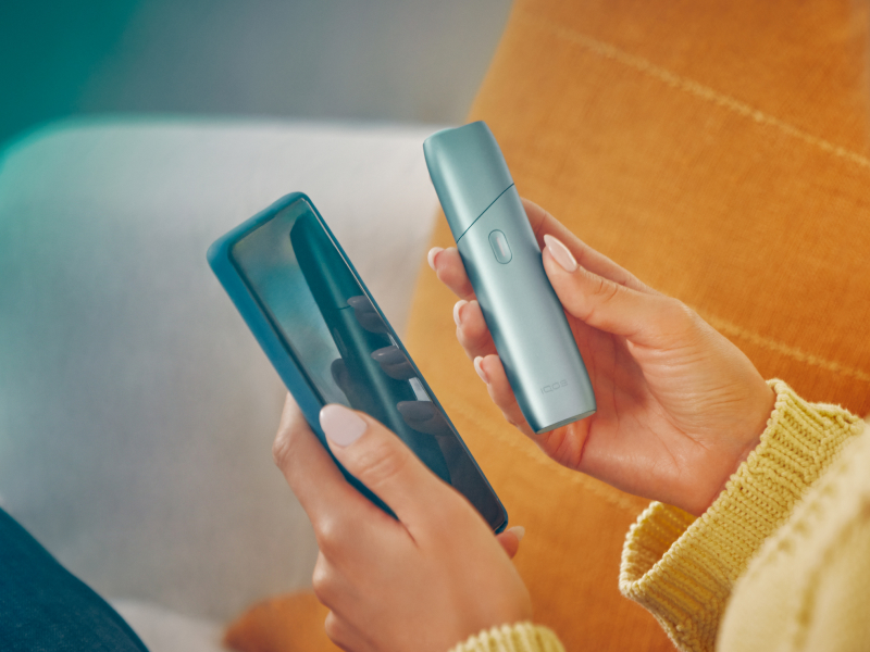 Hands holding a mobile phone and IQOS Originals One device, downloading the app to get the most of the heated tobacco device.