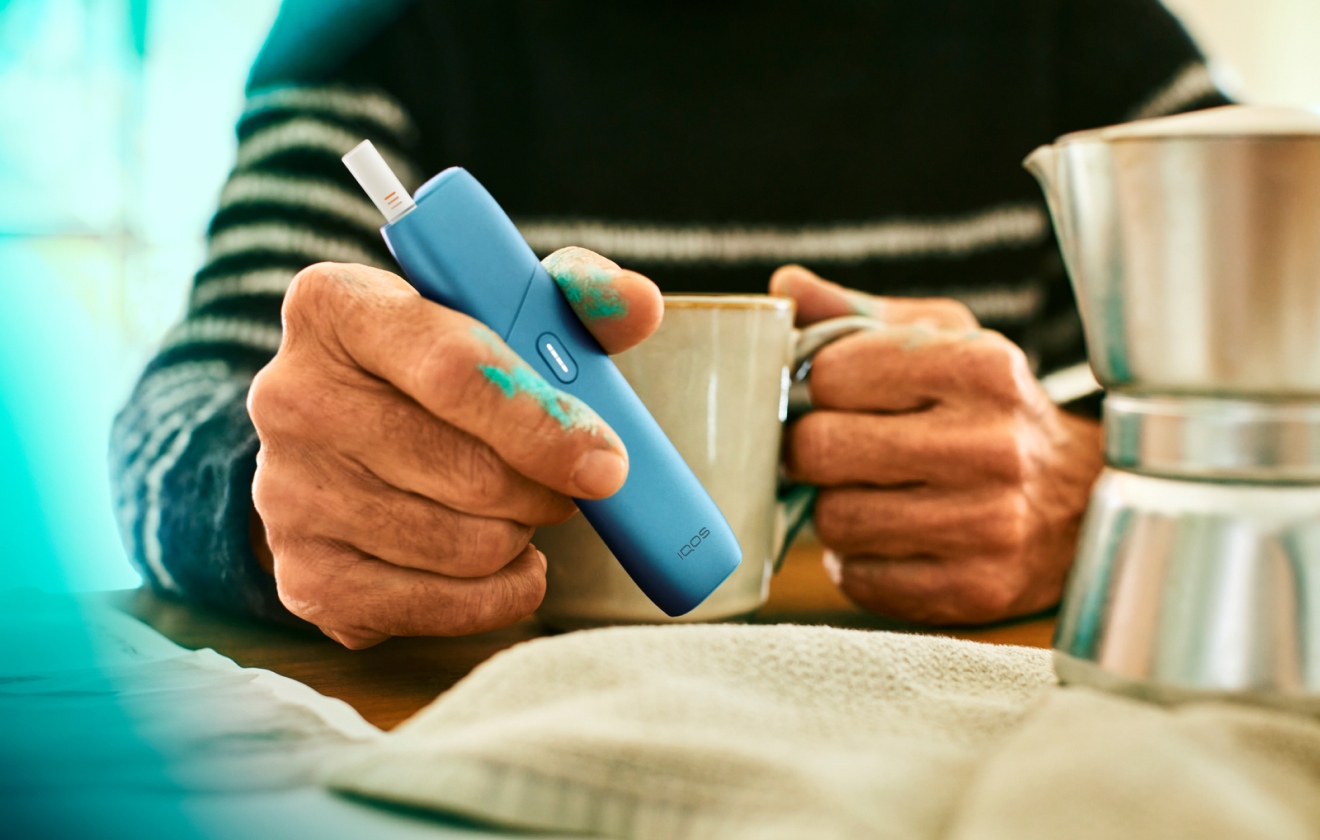 Hands holding turquoise IQOS Originals ONE heated tobacco device and a coffee cup on a table.  